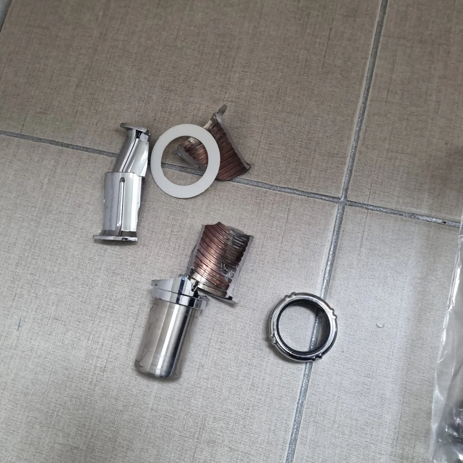 바닥 타일 위에 나사산이 있는 부품 조각 두 개, 흰 와셔, 크롬 고정 링, 마개 부속이 흩어져 있다