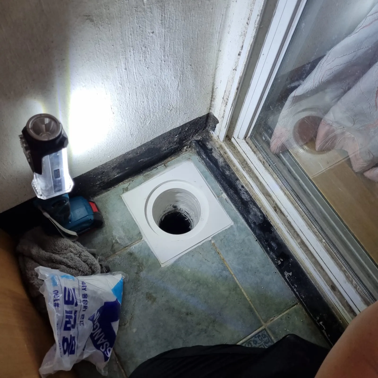 베란다 구석 타일 바닥에 흰색 사각 홈통 뚜껑이 놓여 있고, 가운데 둥근 구멍 안쪽은 어둡다. 왼쪽에는 작업등이 세워져 있다